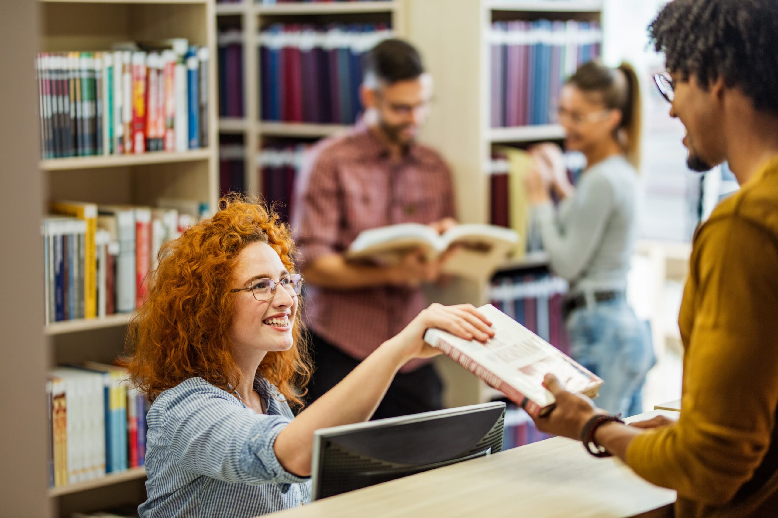 A young woman sits behind a desk and hands a young man a book