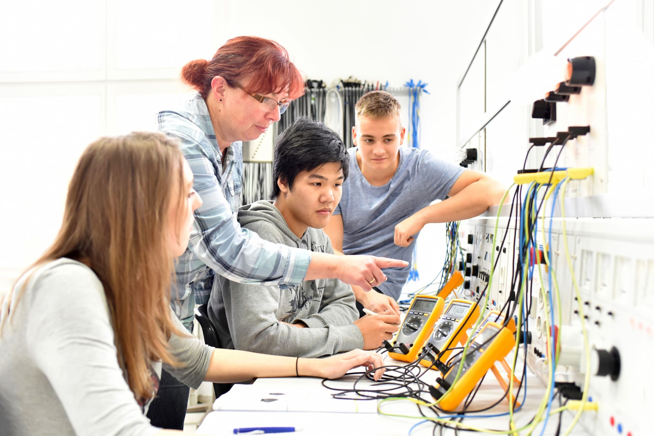 Three teenagers carry out an experiment on a wall with lots of cables and are guided by an adult woman