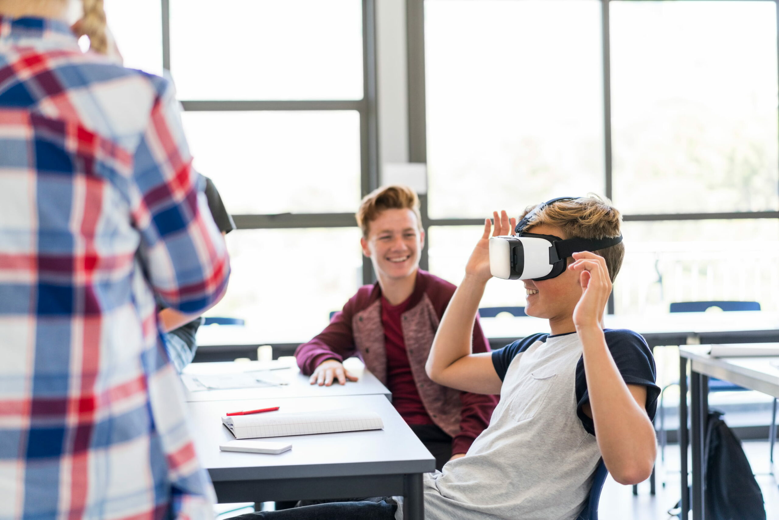 Two boys sit next to each other, one of them wearing VR goggles and testing