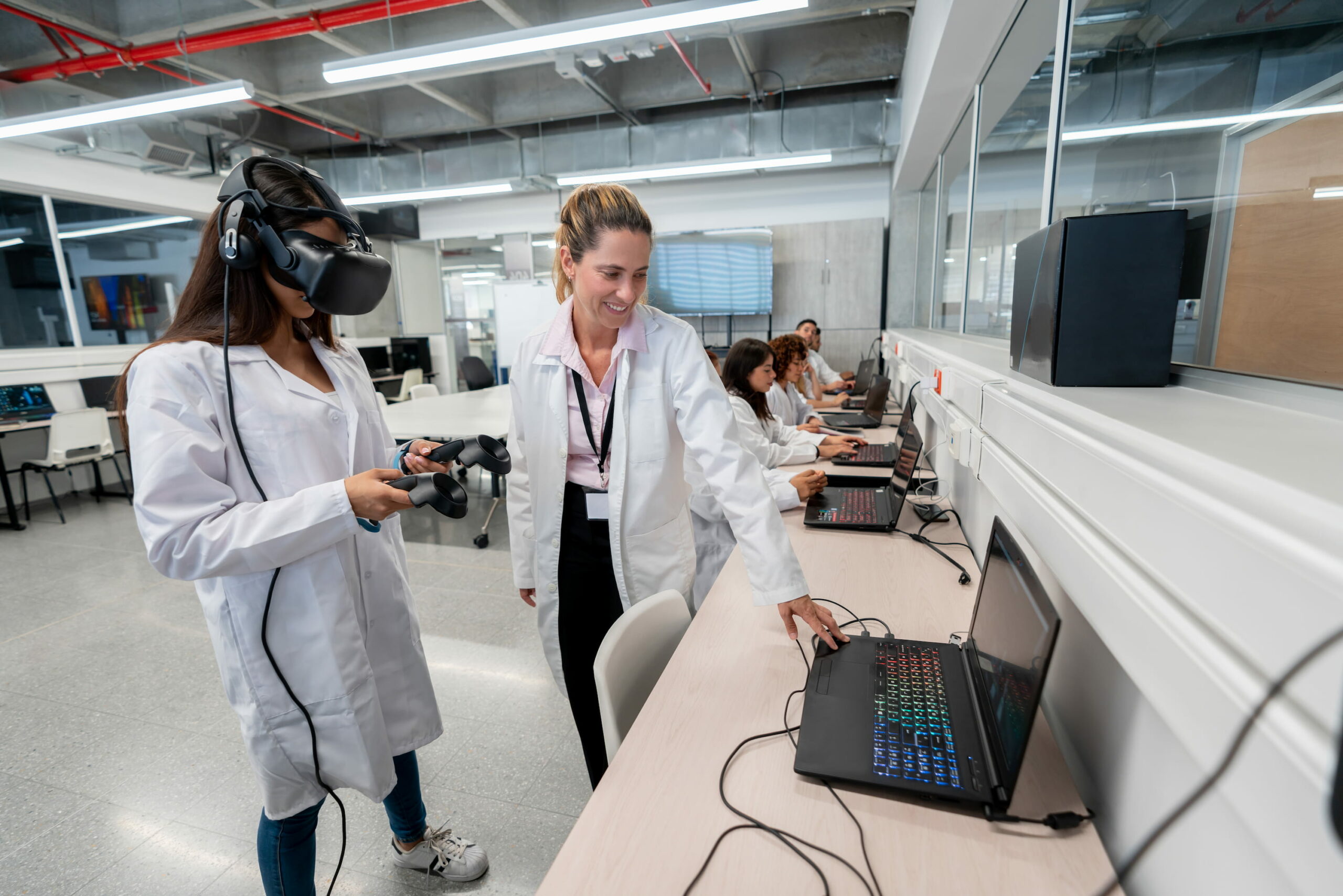 Two women in lab coats stand in front of a desk with a laptop on it, one of them wearing VR glasses and testing them